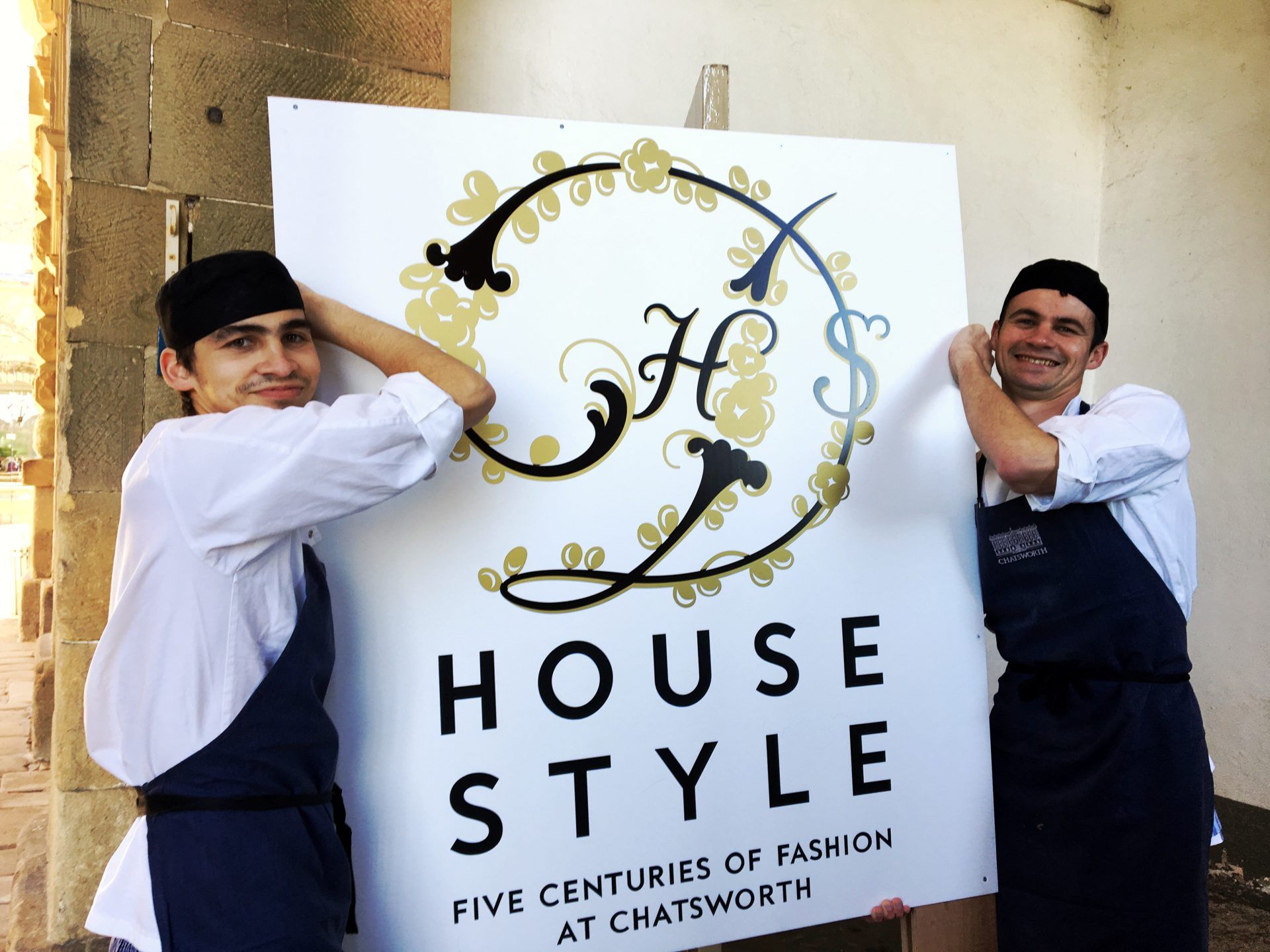 Two of the cafe staff, men in white with black hats and aprons are carrying a sign for the House Style Exhibition