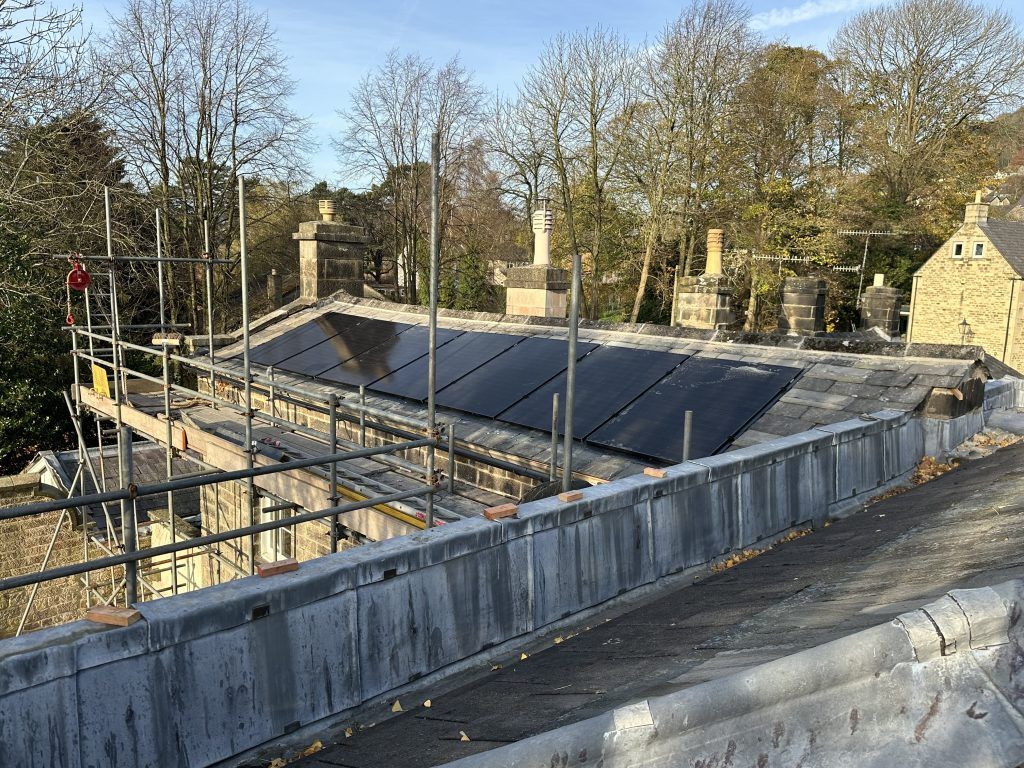 A complex slate tiled roof line showing one section with 7 solar panels and a parapet wall in the foreground all surrounded with scaffolding